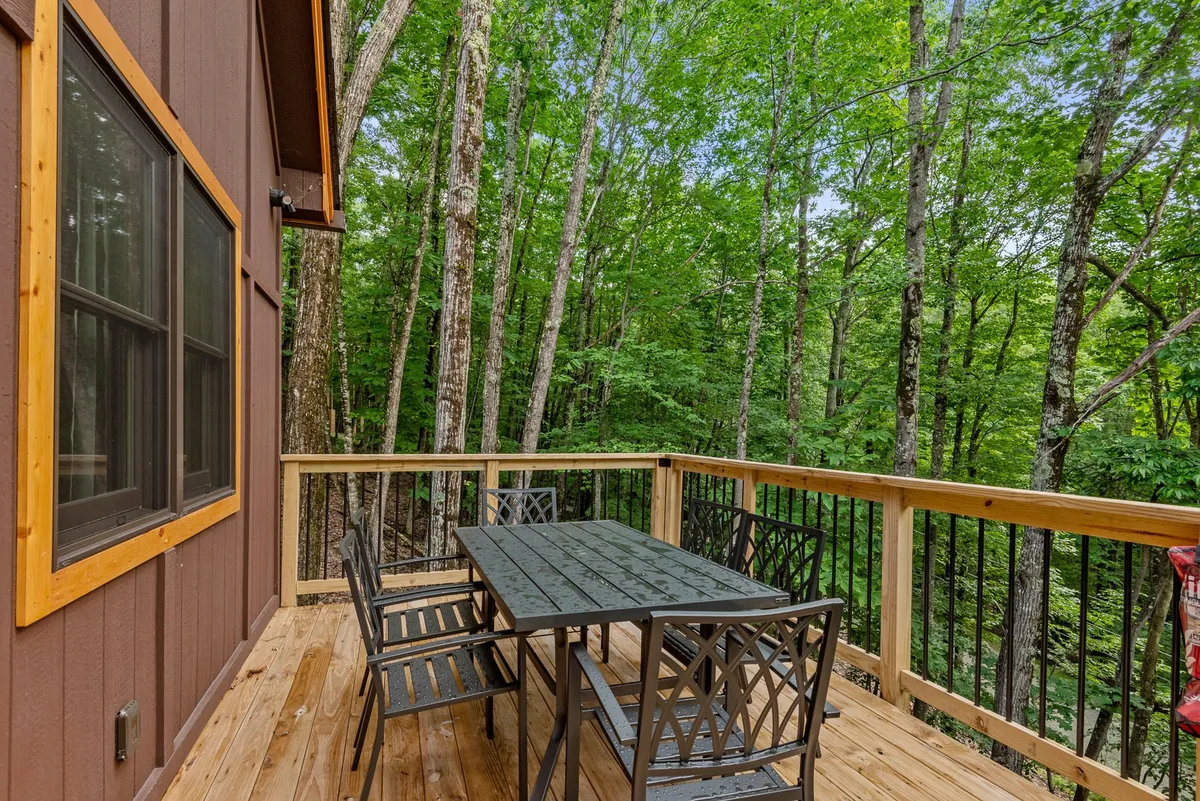 A relaxed deck dining area nestled among trees, with a prominent dining table and railing providing an inviting spot for outdoor meals or coffee. The view is enveloped by nature, suggesting privacy and immersion in the landscape.