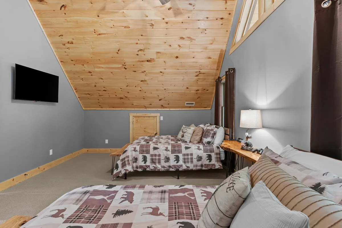 This image features a warmly lit bedroom with a plaid-patterned king bed, wooden bedside table, ceiling fan, and a wall-mounted TV. Large windows let in natural light and offer a view of the surrounding forest, creating a serene retreat atmosphere. The bedding and furnishings evoke coziness and a rustic mountain vibe.