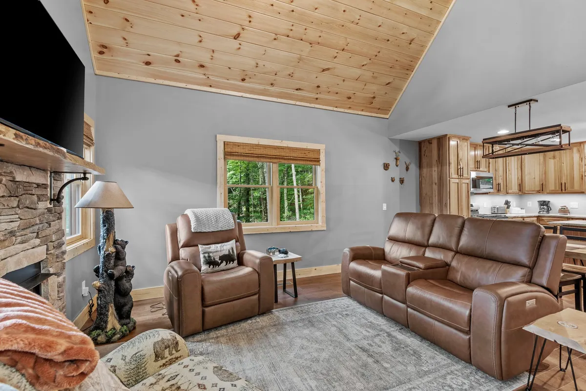 This image showcases a spacious living area with a prominent stone fireplace, comfortable seating arranged to encourage conversation, and a large window offering a forest view. The wooden ceiling and décor elements reflect a mountain-inspired aesthetic, while the abundance of natural light enhances the warm, inviting atmosphere.