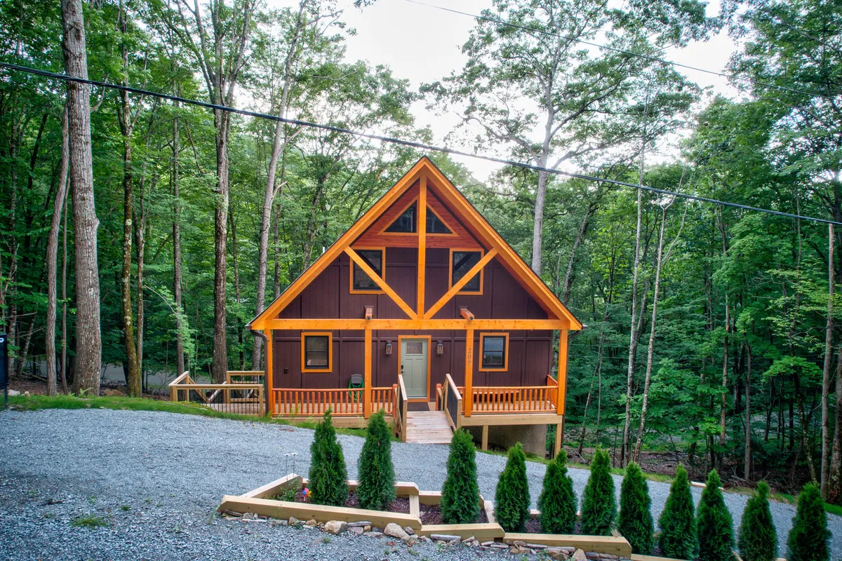 A cozy A-frame cabin with a wooden deck and green landscaping, framed by inviting natural surroundings. The structure’s welcoming design and the deck’s vantage point evoke evenings spent watching stars or listening to the peaceful sounds of nature.