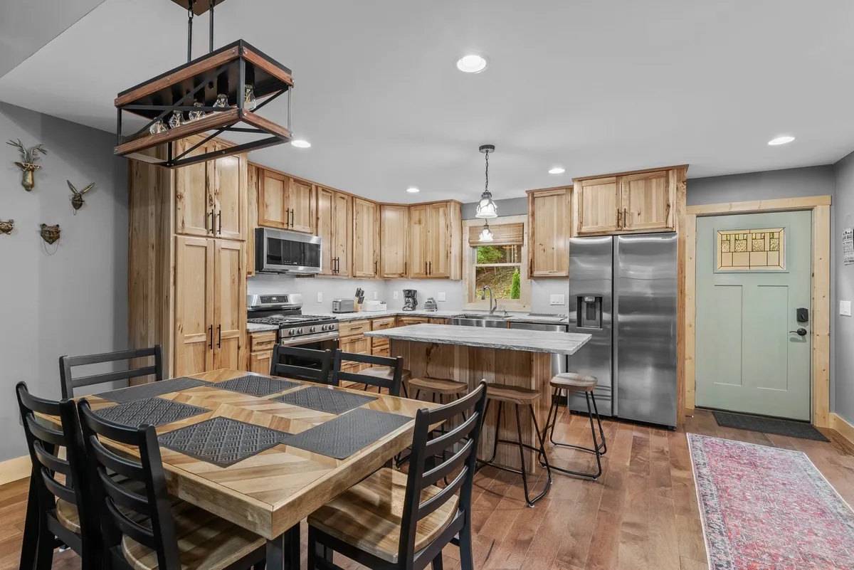 This image showcases the heart of the home, with a warm, inviting view of both the kitchen and adjacent dining table. The kitchen island with seating for three is prominently visible, alongside stainless steel appliances and custom wood cabinetry. The dining table for six is set nearby, offering a clear sense of the communal atmosphere described in the section.