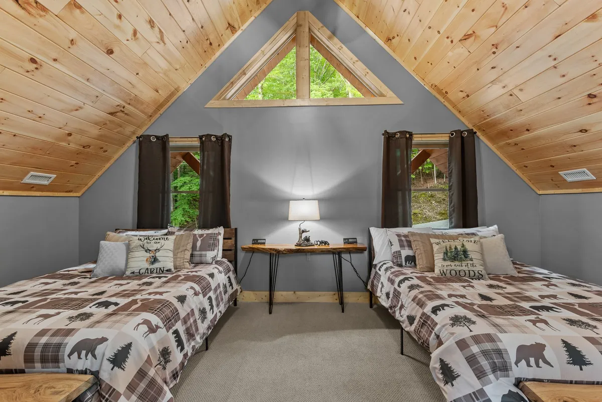 This photo showcases an upstairs bedroom with two beds adorned in patterned quilts, a small wooden table with a lamp between them, and a large triangular window that frames views of the outdoors. The space feels light-filled, comfortable, and designed for relaxation or family gatherings.