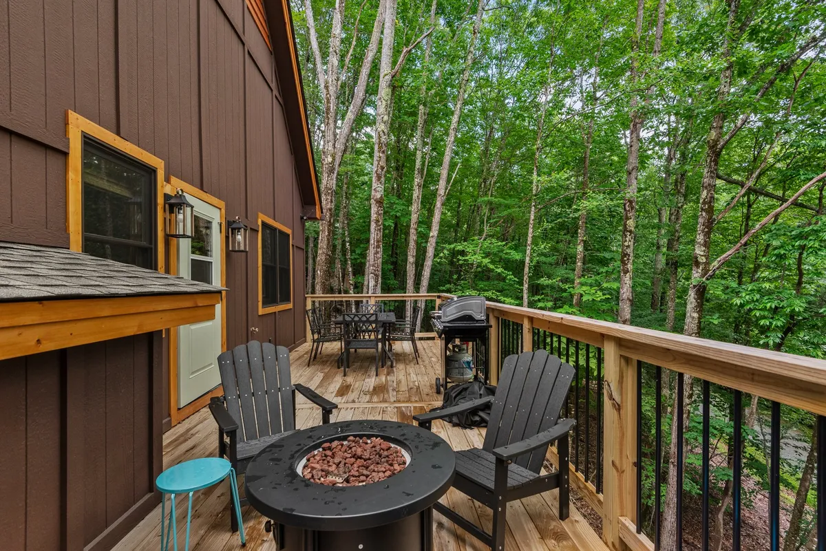 A cozy, pet-friendly wooden deck with a fire pit, adirondack chairs, grill, and forested backdrop. The setup invites guests to gather around the fire, grill, or simply unwind while enjoying the sounds of nature.