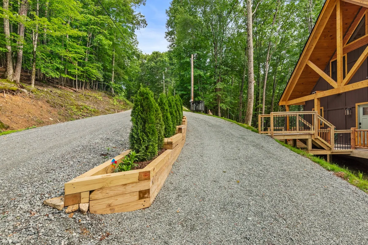 The cabin is viewed from the gravel driveway, framed by mature trees and a welcoming front porch. The exterior shot gives context to the cabin's setting, hinting at the group’s arrival, morning walks, or planning outdoor adventures in the surrounding landscape.