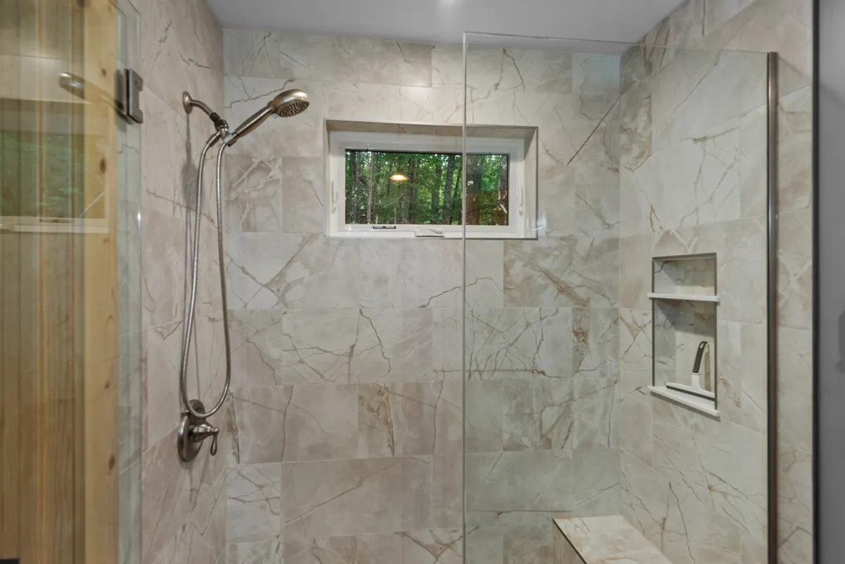 The image focuses on a luxurious bathroom featuring a glass-enclosed walk-in shower with marble tile, a built-in shelf, window for natural light, and modern fixtures. The design suggests spa-like comfort and attention to detail.