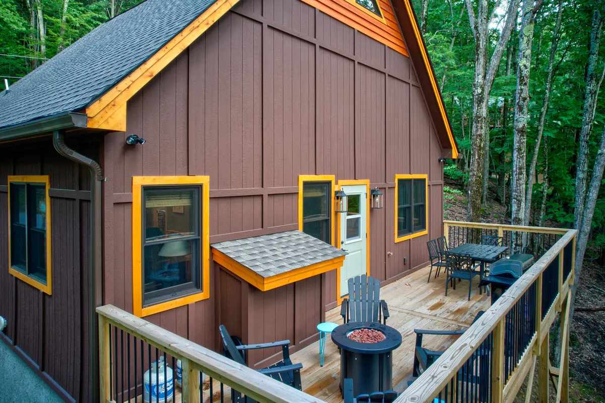 This image features a welcoming deck seating area accented by detailed window frames and vibrant exterior colors, all surrounded by lush trees. The cozy setup suggests a perfect space for relaxing outdoors, sharing a meal, or enjoying the sights and sounds of nature with a pet.